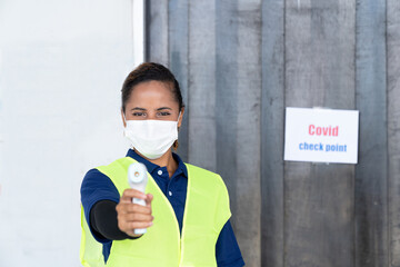 Fototapeta na wymiar Woman worker wearing face mask using infrared forehead thermometer in front of the warehouse at the factory. Factory industry worker operating concept.