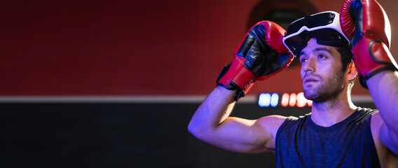 Young caucasian man wearing red boxing gloves and using glasses of virtual reality in sport gym.Smartphone using with VR headset,future technology concept.Man using VR glasses in colorful neon lights.