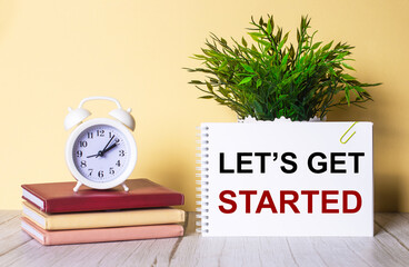 LET IS GET STARTED is written in a notebook next to a green plant and a white alarm clock, which stands on colorful diaries.