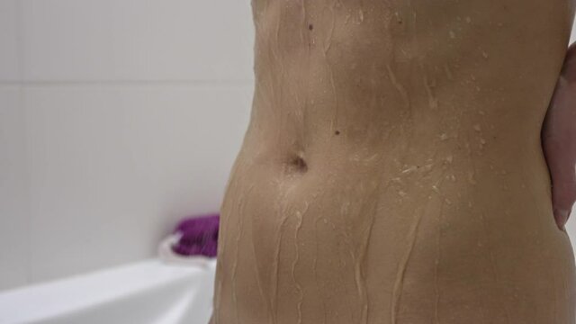 Water flows down the slim belly of a young woman while she is showering.