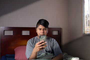 Latin teen checking his cell phone upon waking up in his bed in the morning