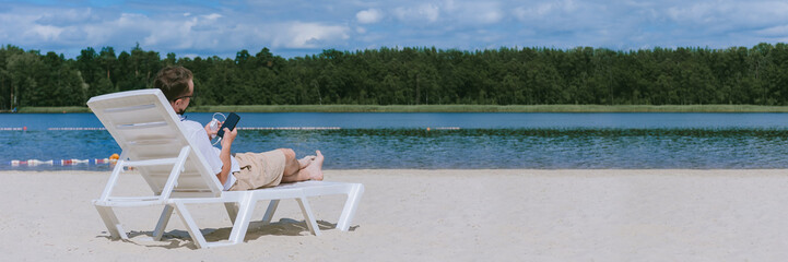 Banner, a man lies on a sun lounger and charges a smartphone from a Power Bank on the beach. Against the backdrop of sand, water and forest.