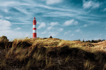 Tall red and white striped lighthouse on sand dunes in evening sunlight. Blue sky with white clouds.