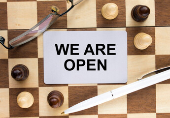 Business card with text We Are Open on a chess board with pen and eyeglasses