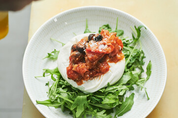 chef pouring grated parmesan over tomato concasse and burrata with rucola inside a restaurant kitchen