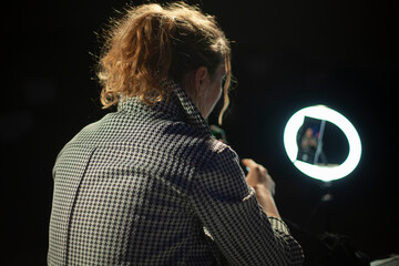 Fototapeta premium The girl removes the blog. The girl uses a round lamp to illuminate her face. Shooting a video blog with artificial light. A woman in a jacket and with long hair tied in a ponytail.