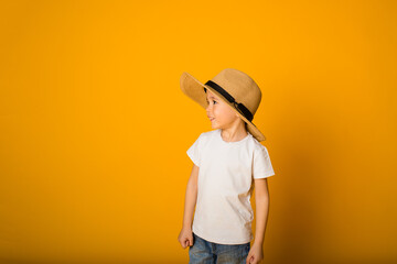 small boy in a white T-shirt and straw hat looks away on a yellow background with space for text