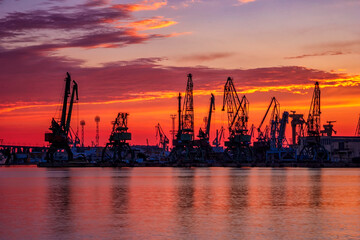 Obraz premium Silhouettes of port cranes at stunning red sunset. Cargo ship terminal at the twilight scene.