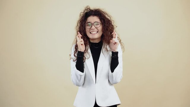 Successful young woman with curly hair, with crossed fingers makes a wish. Young hipster in gray jacket and white shirt, with glasses posing isolated on beige background in the studio.