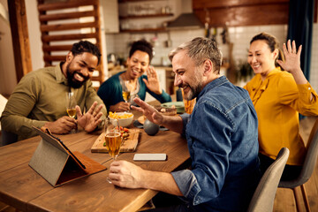 Group of happy friends waving during video call while drinking wine at dining table.