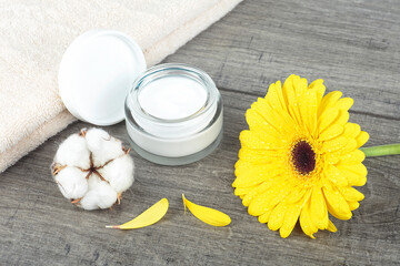 Face cream jar on wooden background with yellow gerbera, soft face towel and cotton flower.