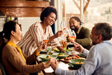 Multi-ethnic group of cheerful friends toasting with wine during meal in dining room.
