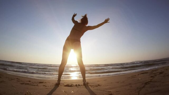 Young fit woman cartwheels at beach sand against sunset, slow motion