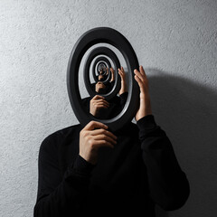 Enigmatic surrealistic optical illusion, young man holding round frame on textured grey background....
