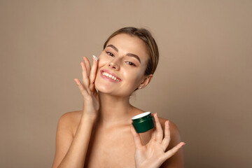 Young woman applies moisturizer to her face and smiles. Skin care concept