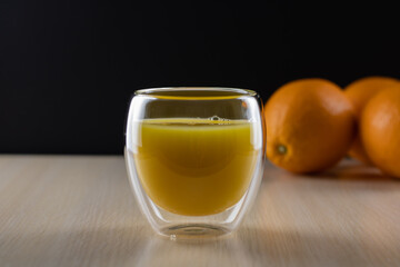 A glass with orange juice in the foreground on a black background. Whole oranges in the background.