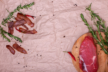 Jerky meat and raw beef on brown craft paper background with copy space. Ingredients for cooking meat snacks. Rosemary, red pepper and peppercorns. Dried meat. Top view