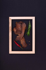 Jerky snacks, red papper and rosemary inside of wooden frame on black background. Creative concept with dried spiced meat for beer