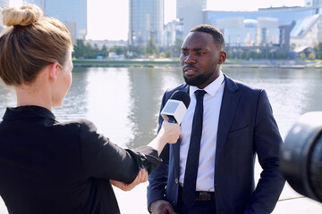Young African entrepreneur standing in front of female reporter or journalist with microphone...