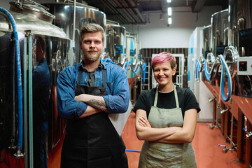 Two happy middle aged workers of beer production factory in aprons crossing arms by chest while standing in front of camera