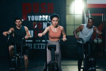 Multi ethnic group of young athletes training on stationary bike cardio machine building muscle in fitness gym.