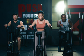 Multi ethnic group of young athletes training on stationary bike cardio machine building muscle in fitness gym.