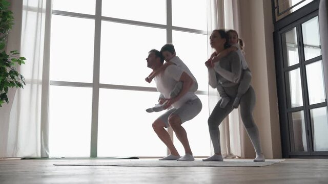 Full length view of active parents doing squats or situps with little kids piggybacking. Father and mother working out together with children at home
