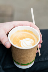 Recyclable coffee cup held in a hand of caucasian man along with wooden stiring stick, tasty caffeine drink with cream heart shape in paper single use cup, eco friendly approach for takeaway drinks