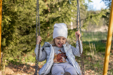 A girl in a cap and vest on a swing. Blonde baby in a good mood. Little girl having fun on a chain swing. spring in the park