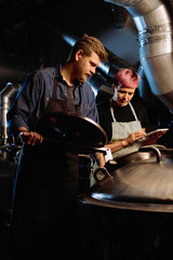 Young woman with tablet scrolling through list of beer processing stages while standing next to her male colleague opening lid of cistern