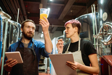 Young bearded brewer in apron looking at beer in glass while standing next to his female colleague...