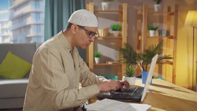 Muslim in a skullcap works at home on a laptop