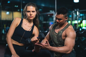 Caucasian young woman in sportswear lifting dumbbell  with trainer care for safety in fitness club or gym.