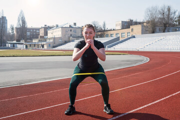  Young sportive woman trains legs with the help of fitness rubber bands on the red track.