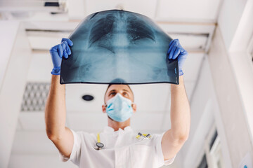 Doctor with face mask and rubber gloves standing in hospital and looking at x-ray of lungs during...