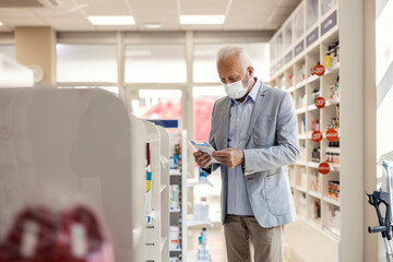 A patient in a pharmacy. An older man with gray hair in an elegant suit holds medication in his hand and reads therapies. He is in a pharmacy lit by daylight. Protective mask against corona virus