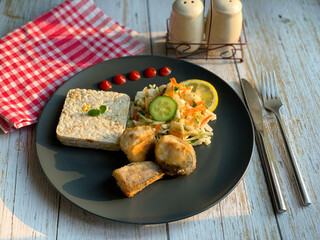 Vegetable cabbage salad and fried fish on a plate. Diet bread for lunch. Cutlery and black plate on the table.