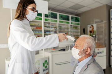 Therapy in a nursing home pharmacy at the time of corona virus. A young pharmacist measures the temperature of an adult man sitting in a chair in an elegant suit. They wear face masks