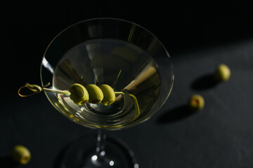 Glass of classic dry martini cocktail with olives on dark stone table against a black background.