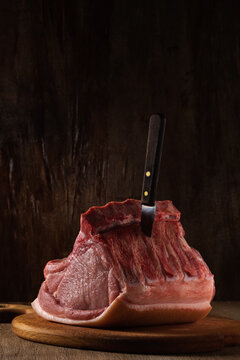 a whole freshly cut piece of raw pork loin lies on a cutting board with a stuck knife a brown wooden backdrop. side view. artistic moody vertical photo in simple rustic style with copy space