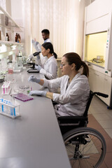 Naklejka premium Disabled scientist sitting at the table and working with samples in test tubes with her colleagues in the background