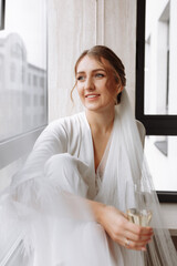 Bride s morning. Bride drinking champagne in the peignoir. young woman is sitting on a large window in a hotel room in bathroom. Beautiful girl in white wedding robe. wedding day