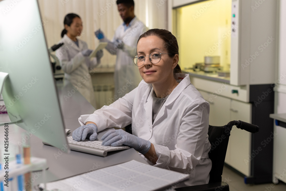 Sticker portrait of female chemist in eyeglasses looking at camera while sitting at the table and working on