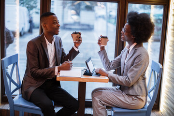 Smiling afro american man and woman drinking fresh coffee at cafe while leading business meeting. Two colleagues in formal wear using modern gadgets for work.