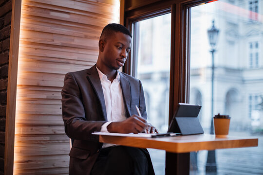 Young african businessman in stylish suit taking notes while looking at tablet screen. Confident man sitting at cafe table and working on distance.