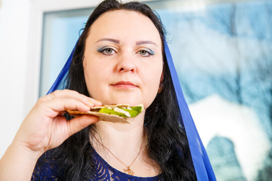 A Jewish woman with her head covered in a blue cape at the Passover Seder table is eating moror hazeret matzah.