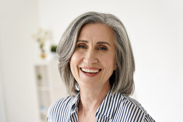 Cheerful satisfied 50s mature woman laughing looking at camera at home. Happy sophisticated classy mid age older gray-haired lady with white teeth dental smile posing for close up headshot portrait.