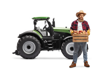 Fototapeta premium Full length portrait of a farmer with a crate full of fresh apples standing in front of a tractor