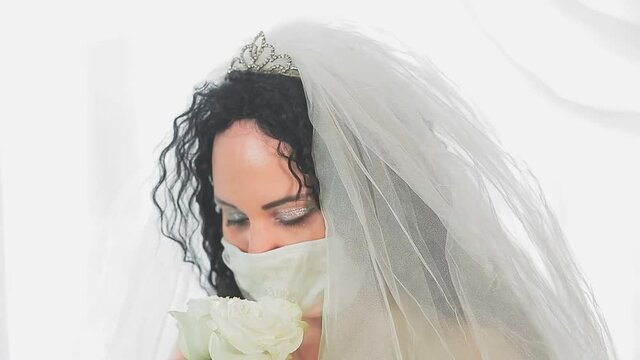 The face of a Jewish bride in a wedding dress and veil in a medical mask in the synagogue hall before the chuppah ceremony with a bouquet of flowers. From blur to sharpness