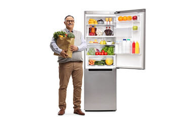 Full length portrait of a mature man with a grocery bag standing next to an open full fridge